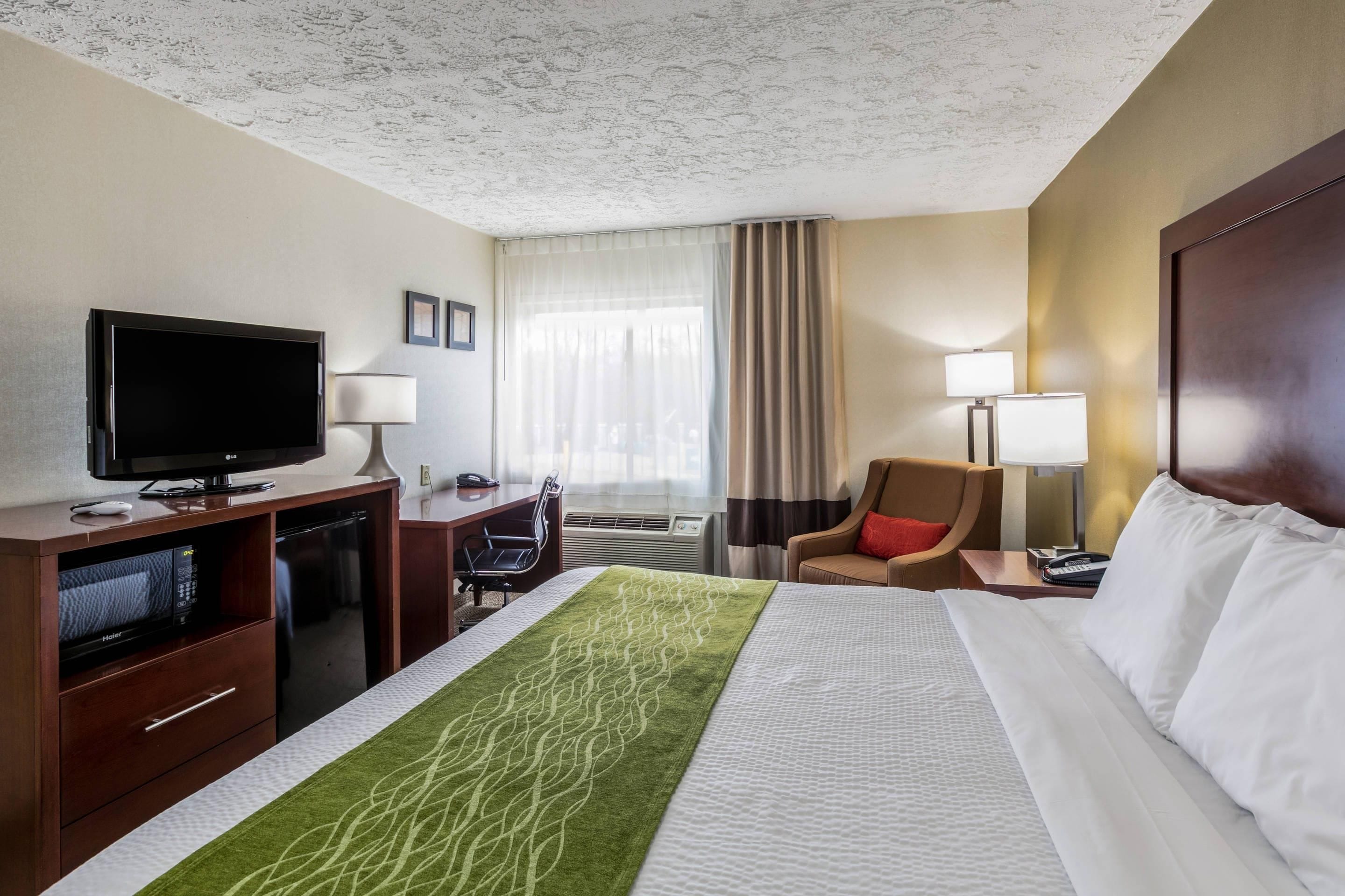 A hotel room with a bed, a desk with a television, a chair, a desk lamp, and a window with curtains.