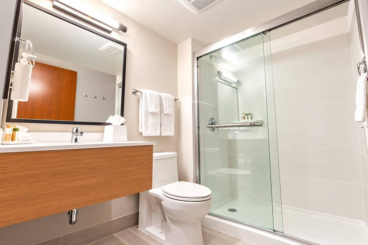 A modern bathroom with a glass-enclosed shower, a wooden vanity, a white toilet, and a large mirror.