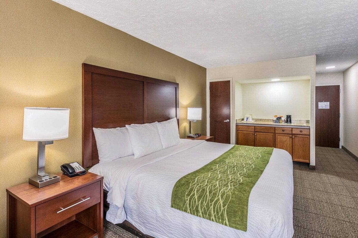 A hotel room with a bed, bedside lamps, a desk with a phone, a mirror, and a kitchenette.