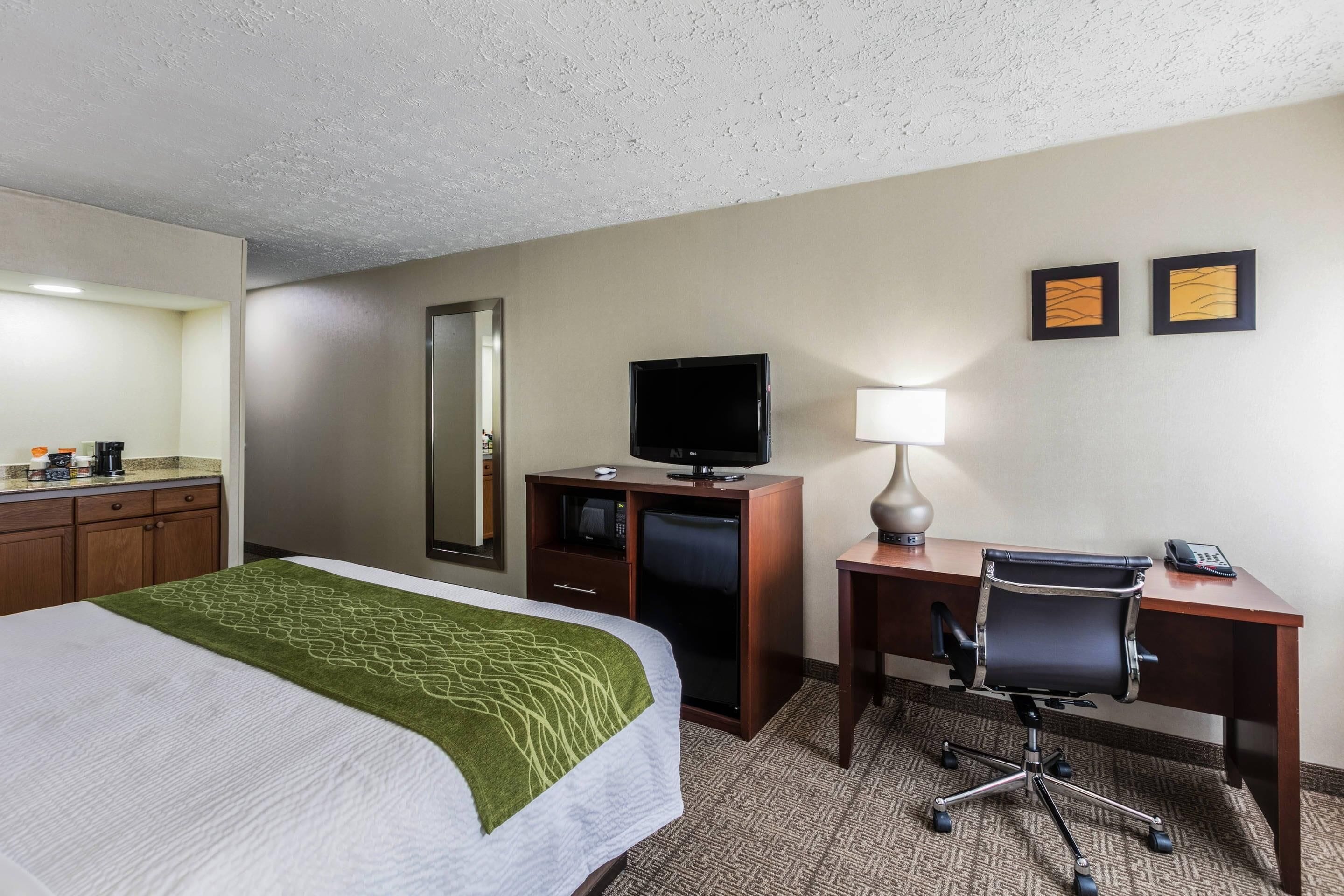 A hotel room with a bed, a desk, a chair, a television, a mirror, and two framed pictures on the wall.
