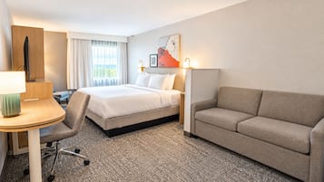 A hotel room with a bed, a sofa, a desk with a lamp, a chair, and a painting on the wall.