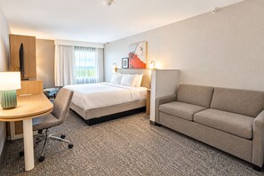 A hotel room with a bed, a sofa, a desk with a lamp, a chair, and a painting on the wall. - Comfort Inn Medford - Long Island (Medford)