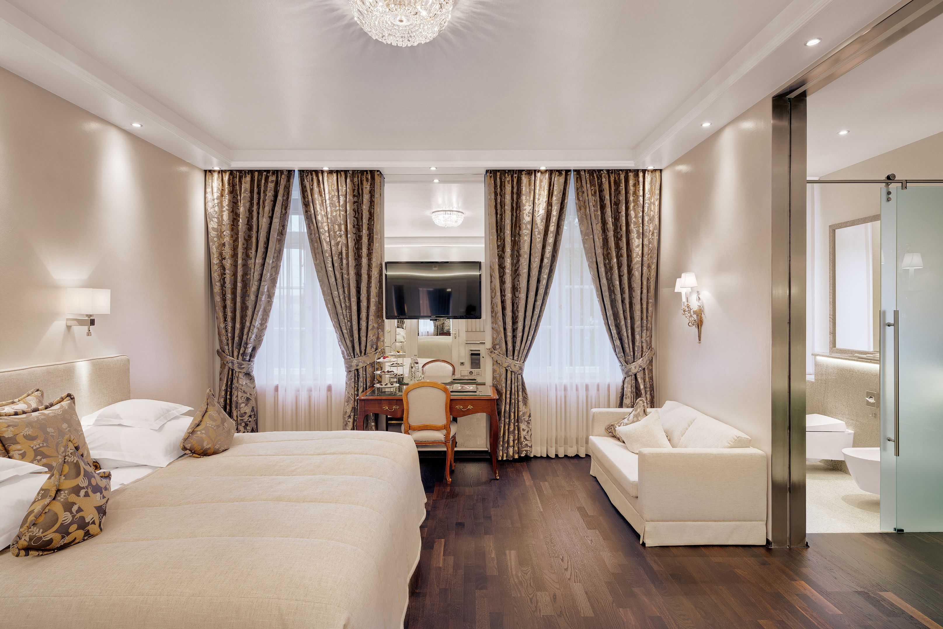 A hotel room with a large bed, a desk with a chair, a sofa, and a bathroom visible through an open door.
