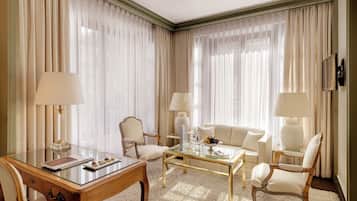 Ein Hotelzimmer mit einem Glastisch, zwei Sesseln, einem Sofa, einem Schreibtisch mit Lampe und transparenten Vorhängen.