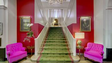 A grand staircase with a green carpet, flanked by two vibrant pink armchairs and a vase of flowers.