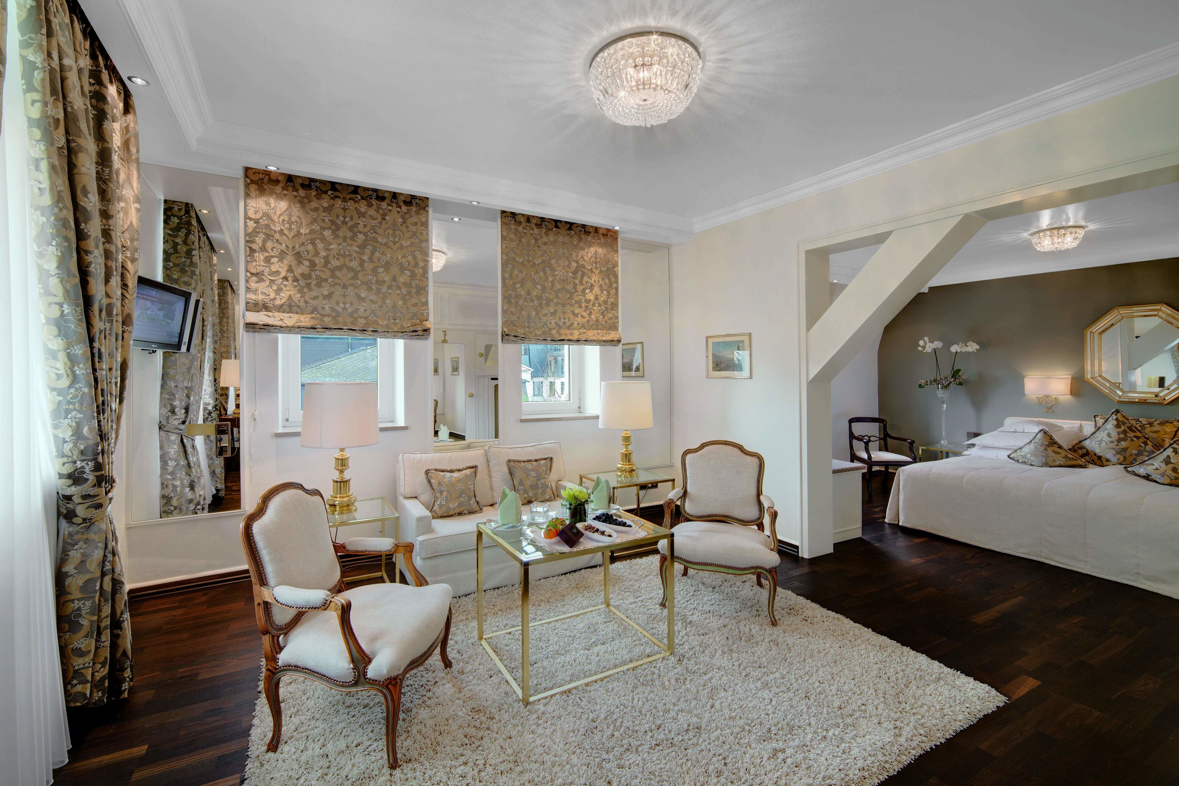 A spacious bedroom with a large bed, a sitting area with chairs, a dining table, and a chandelier.