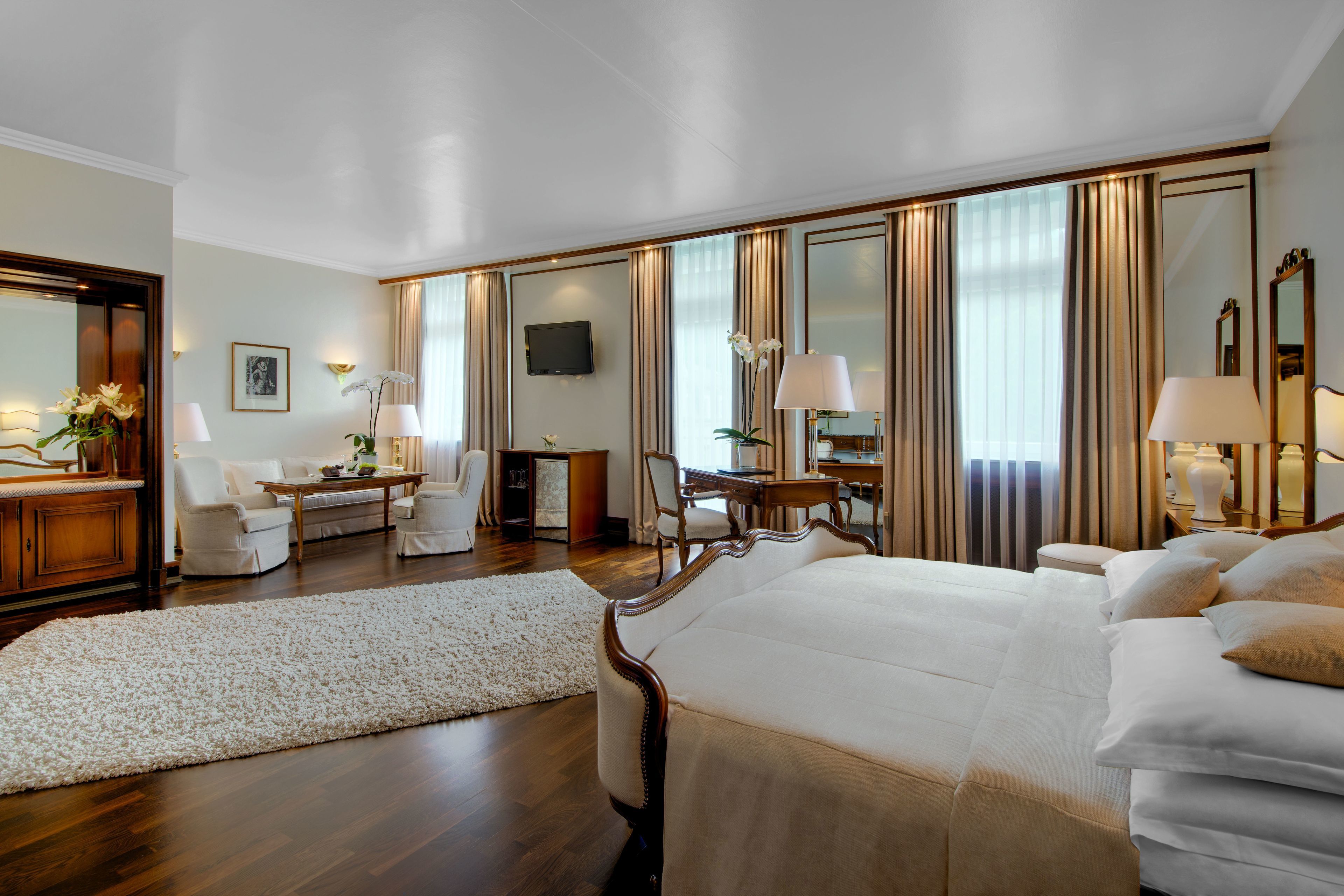 A spacious hotel room with a large bed, a sitting area with a desk and chairs, a television, and a large mirror.