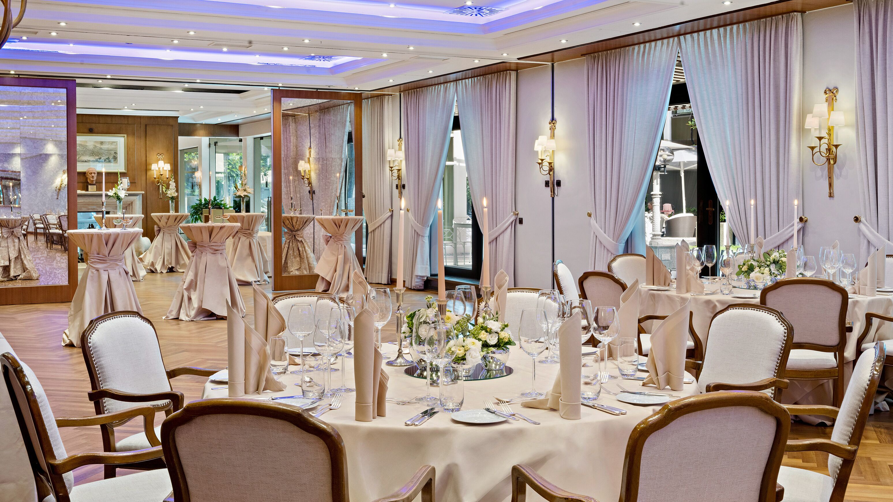 A formal dining room with tables set for a meal, featuring elegant chairs and candle centerpieces.