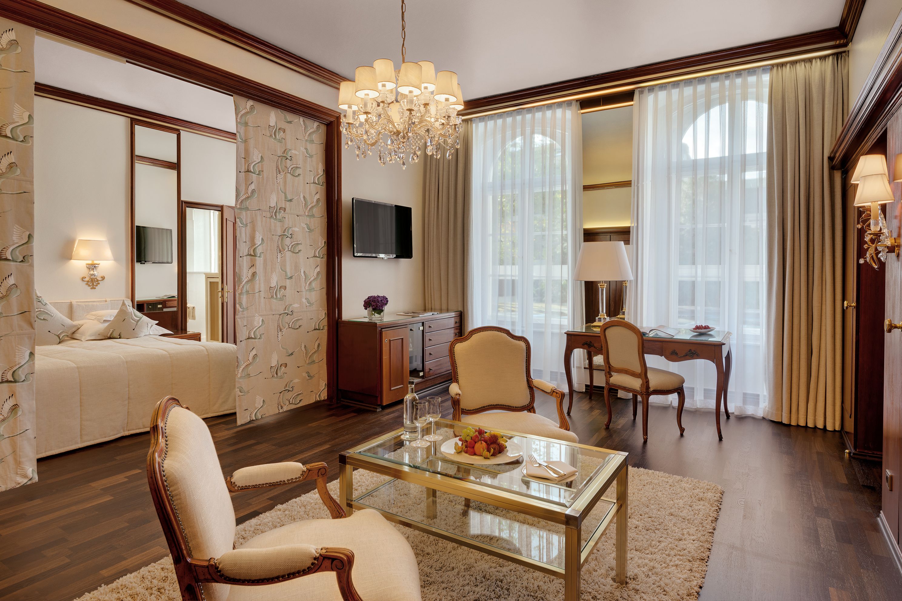 A spacious hotel room with a large bed, a sitting area with a glass coffee table, a dining area with a table and chairs, a television, and a chandelier.