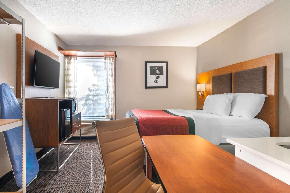 Habitación de hotel con cama, escritorio, silla, televisión y ventana con cortinas.