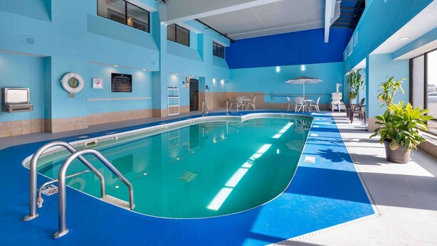 Una piscina interior con agua cristalina, rodeada de paredes y suelo azules. Hay barandillas de metal a lo largo del borde de la piscina y algunas plantas en macetas cerca de ella.