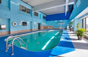An indoor swimming pool with clear water, surrounded by blue walls and a blue floor. There are metal handrails along the pool's edge and a few potted plants near the pool's edge.