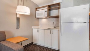 Una kitchenette compacta con microondas, lavamanos y refrigerador. Hay una mesa pequeña y una silla, y las paredes son blancas.