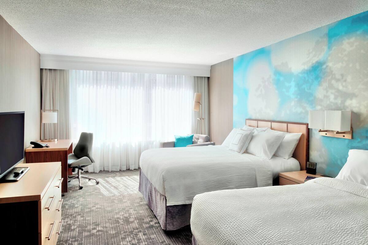 A hotel room with two beds, a desk, a chair, a TV, and a large window with curtains.