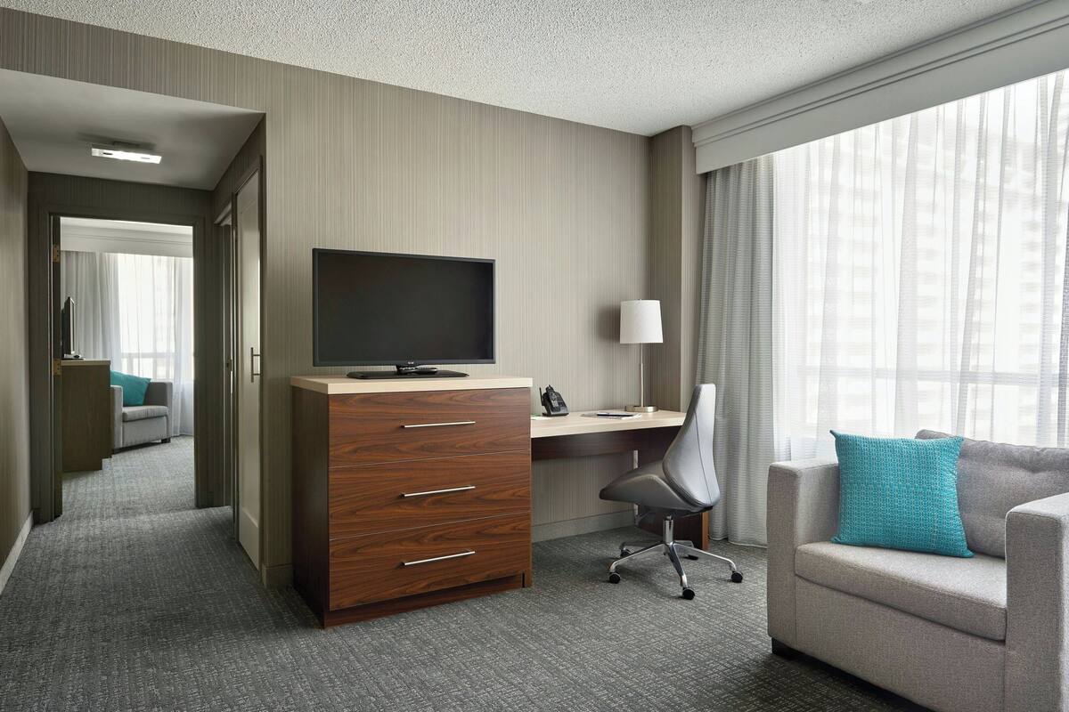 A hotel room with a desk, a television, a chair, a sofa, and a window with curtains.