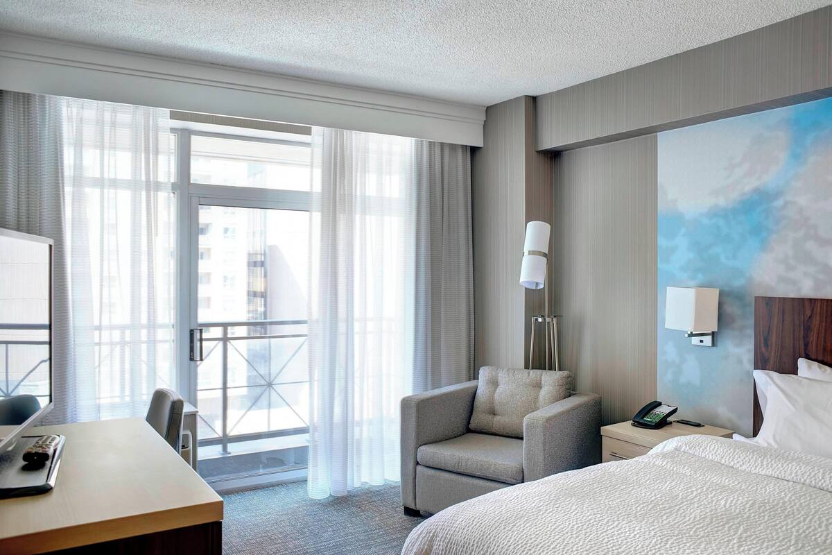 A hotel room with a bed, a desk, a chair, a nightstand with a phone, and a window with curtains.