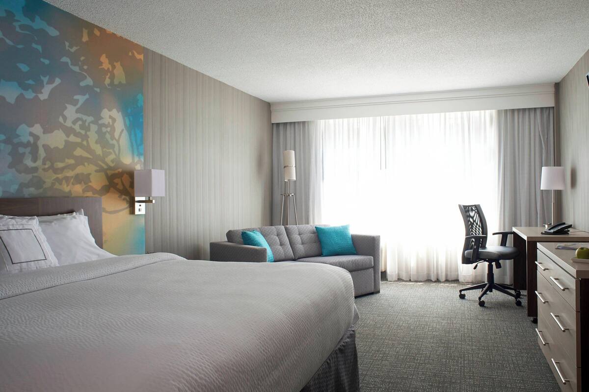 A hotel room with a large bed, a sofa, a desk with a chair, and a large window with curtains.