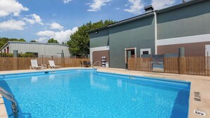 A rectangular swimming pool with clear blue water, surrounded by a tiled deck. There are two white lounge chairs and a small table with a chair. A building with a flat roof and a brick wall is adjacent to the pool.
