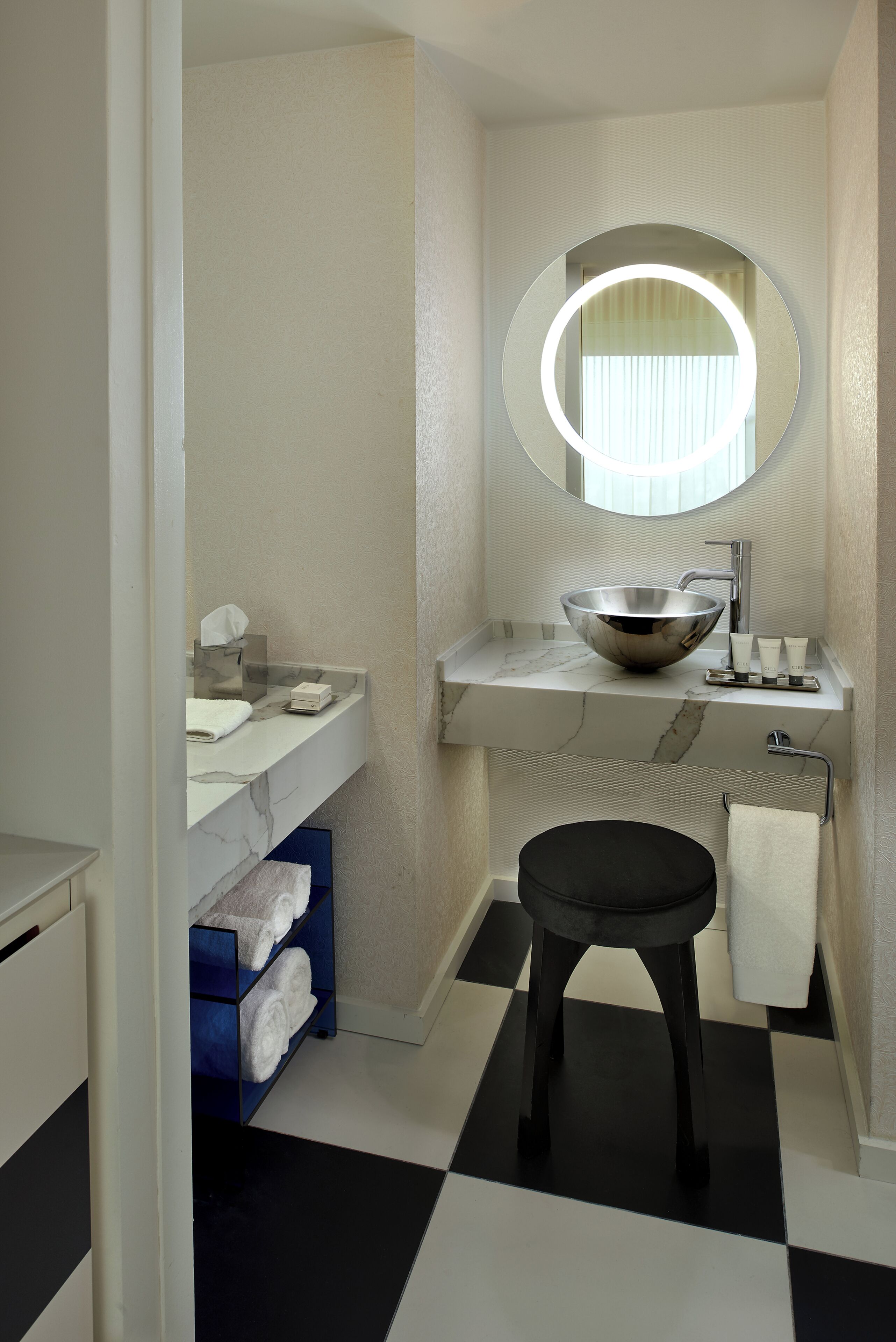 A bathroom with a round mirror, a bowl sink, a towel rack, and a black stool.