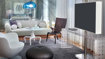 Una sala de estar moderna con un sofá, sillas, una mesa de centro de vidrio, un reposapiés negro y un televisor sobre un soporte transparente.