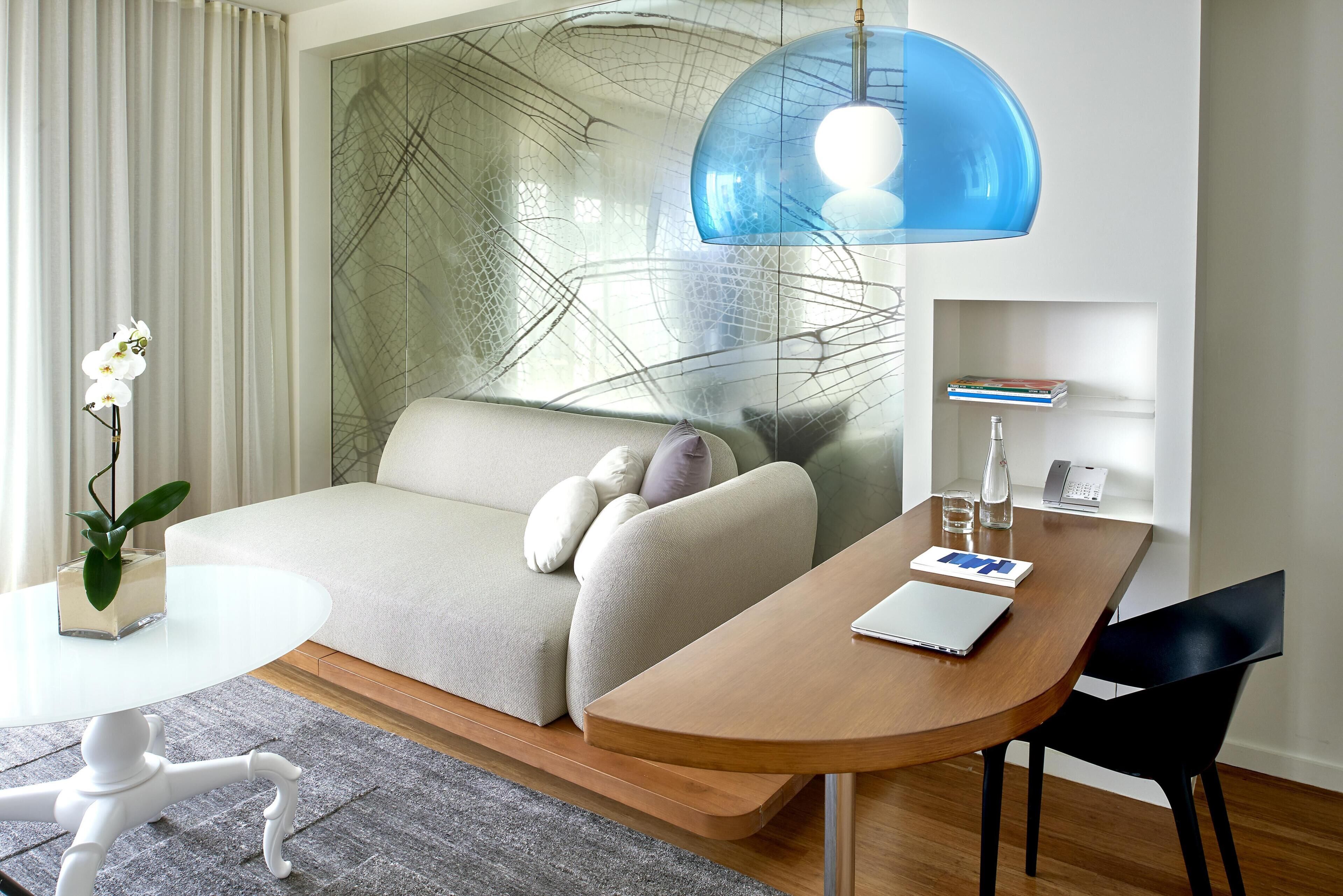 Ett modernt hotellrum med en soffa, ett skrivbord i trä, en bärbar dator, en vas med blommor och en stor, abstrakt väggdekoration.