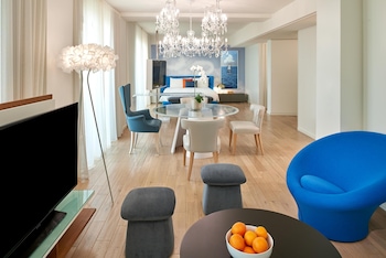 A modern bedroom with a large bed, a TV, a glass dining table with a bowl of fruit, and a decorative chandelier. at The Valorian Los Angeles, Curio Collection by Hilton