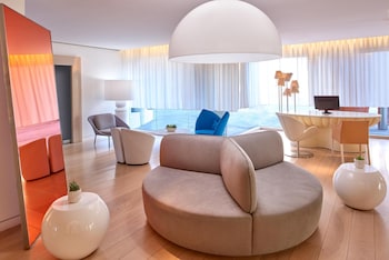 A modern hotel room with a flat-screen TV, a dining area with a glass table, blue chairs, and a chandelier. at The Valorian Los Angeles, Curio Collection by Hilton