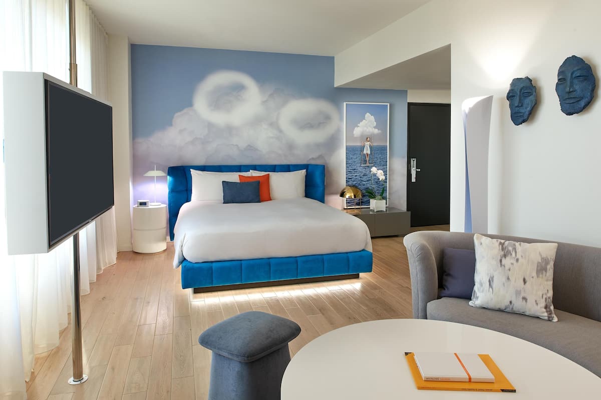 Ein modernes Hotelzimmer mit Bett, Sofa, Fernseher und Wand mit Wolkenmotiv.