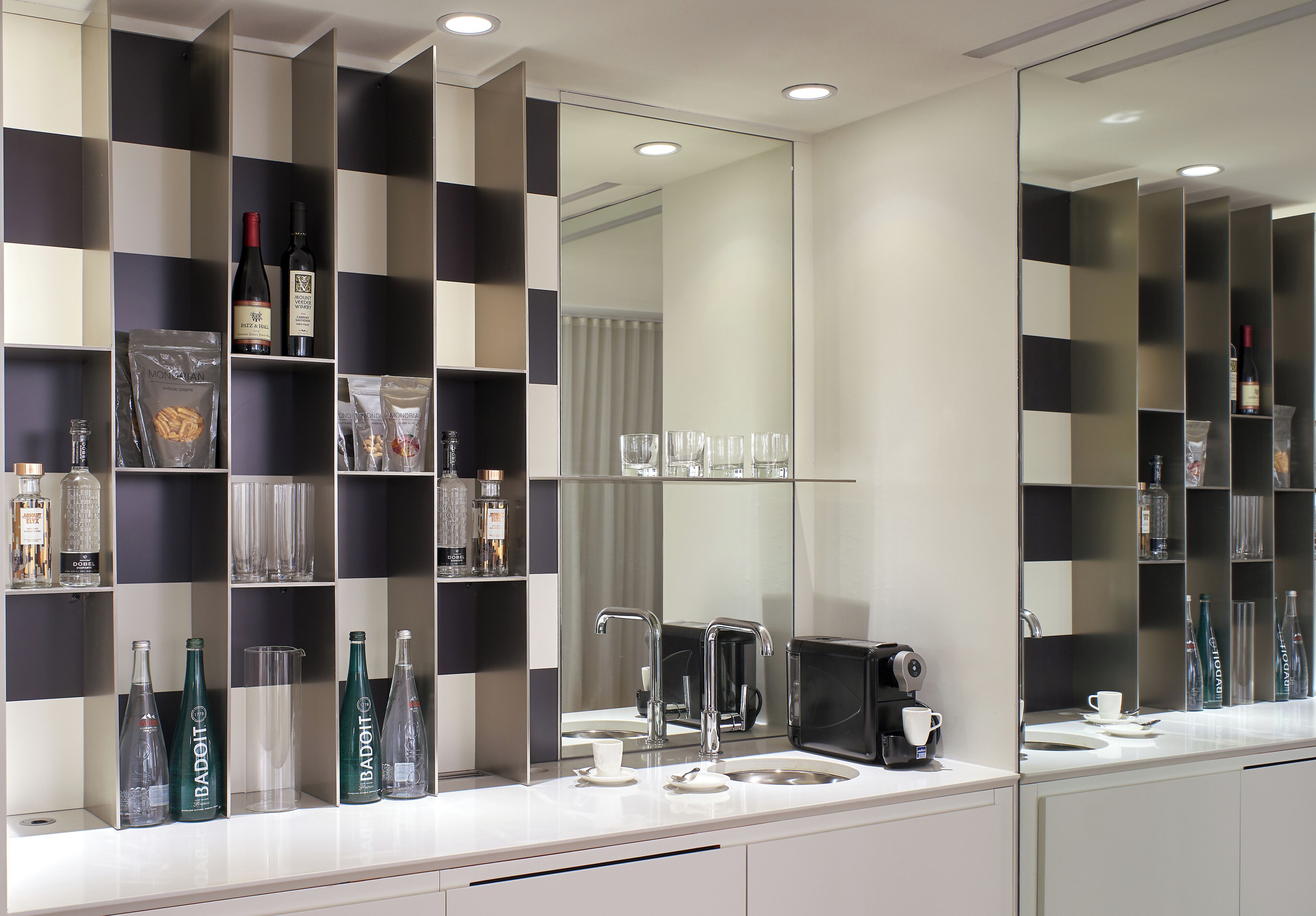 A modern bathroom with a large mirror, a coffee machine, and neatly arranged shelves with bottles and glassware.