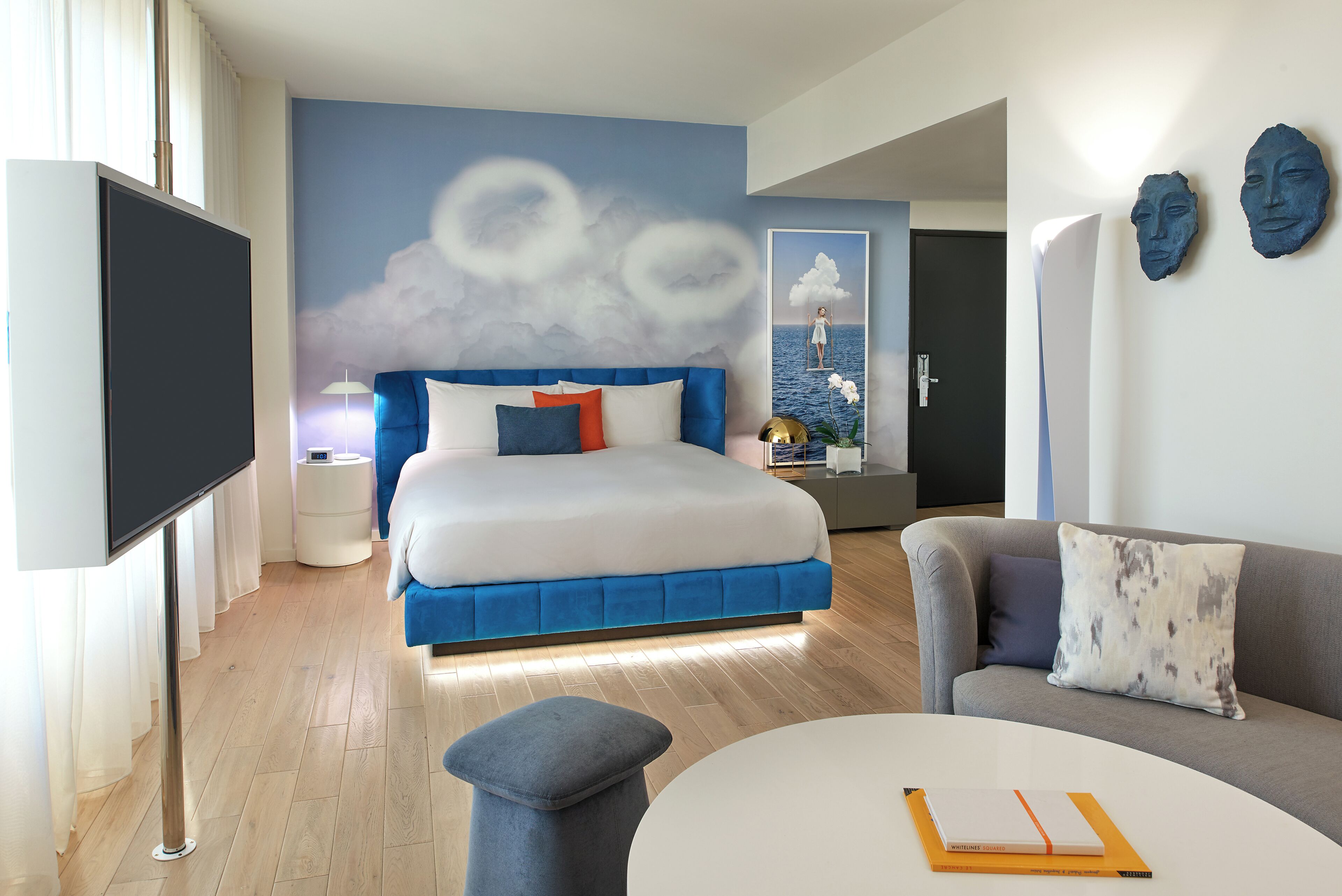 Una habitación de hotel moderna con una cama, un sofá, un televisor y una pared con un mural de nubes.