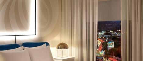 A hotel room with a bed, a nightstand, a lamp, and a view of a cityscape at night.