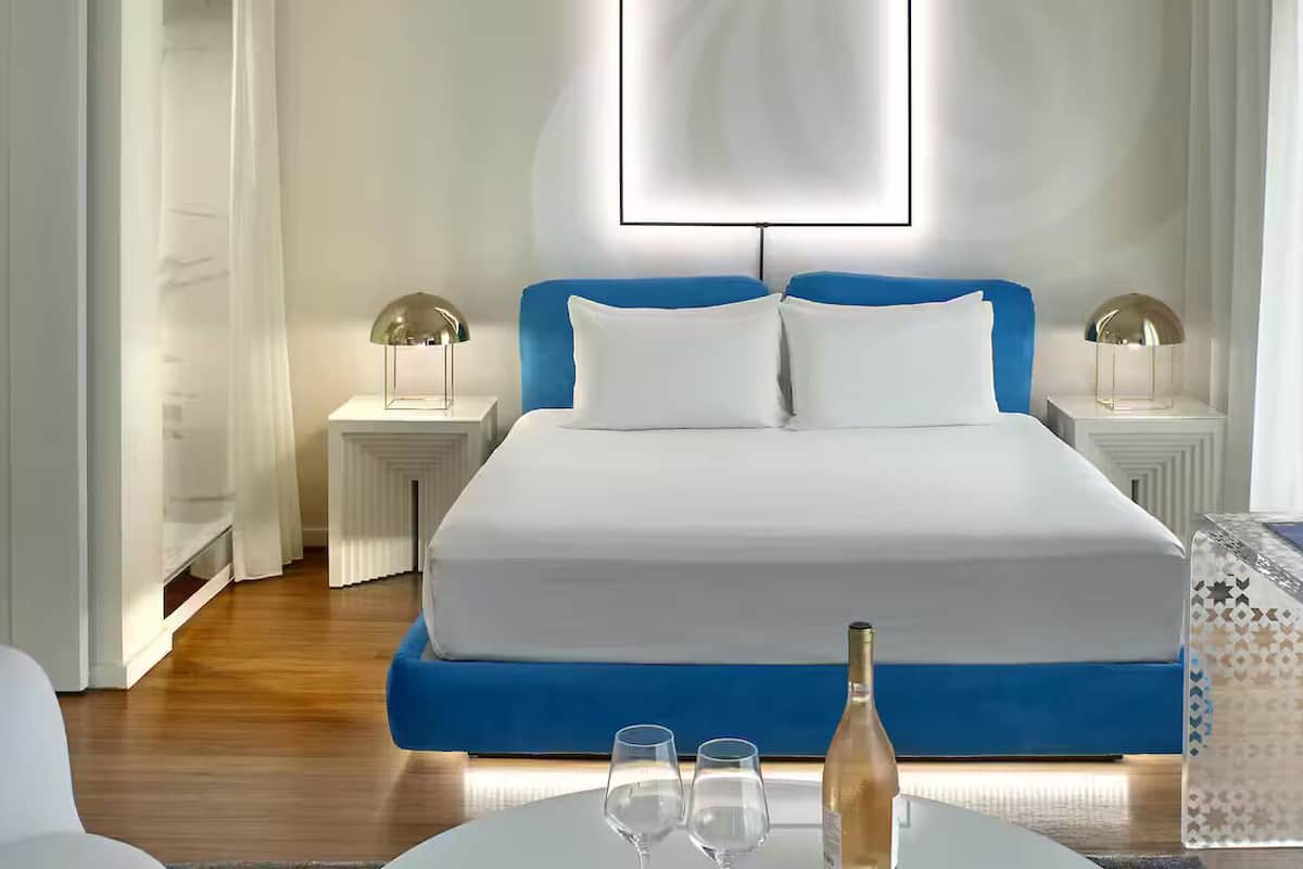 Ein Hotelzimmer mit Bett, Nachttischlampen, einem Glastisch mit Flasche und Gläsern sowie freiem Blick auf den Flur.