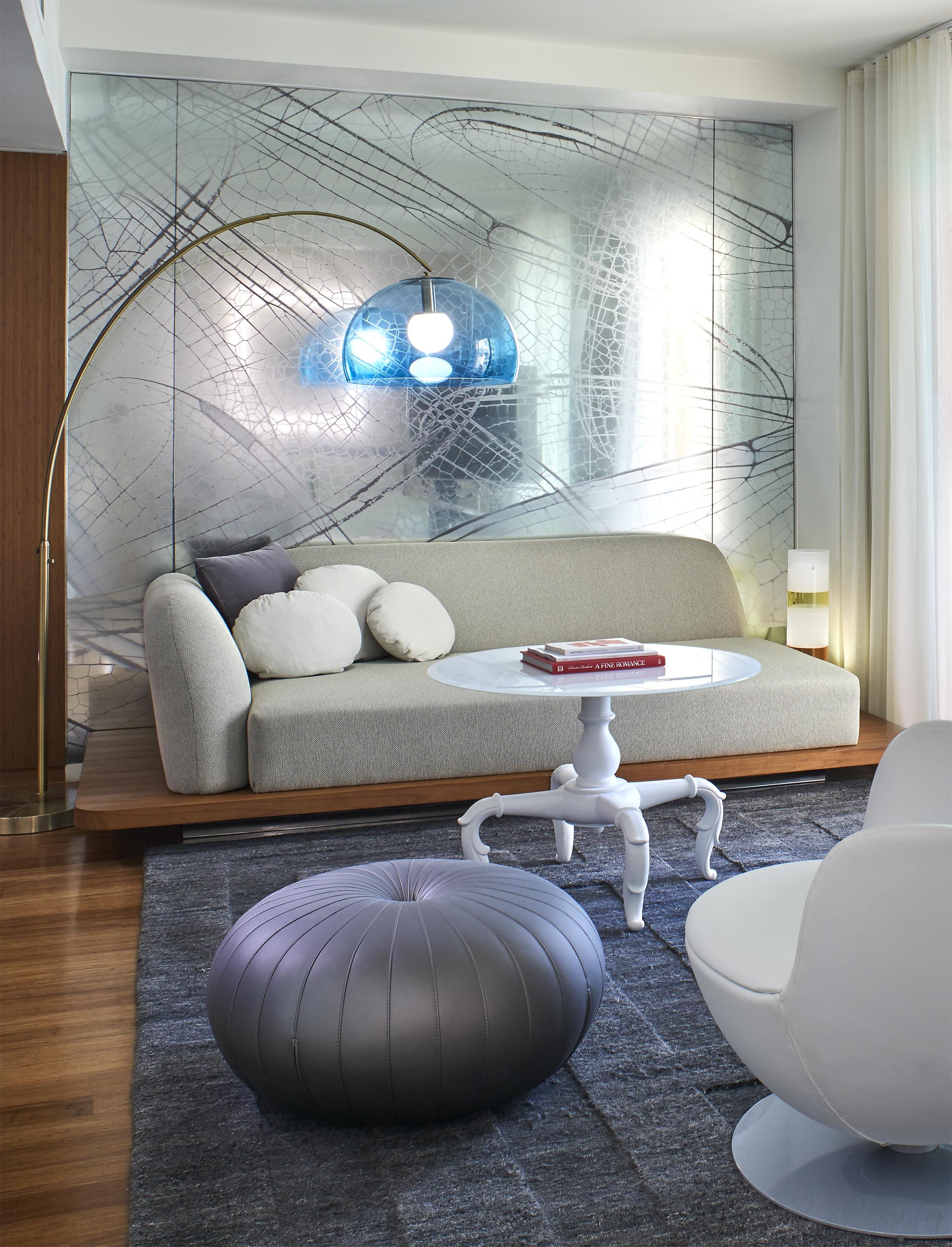 A modern living room with a sofa, ottoman, and chairs, a glass wall with a geometric pattern, and a blue pendant light.