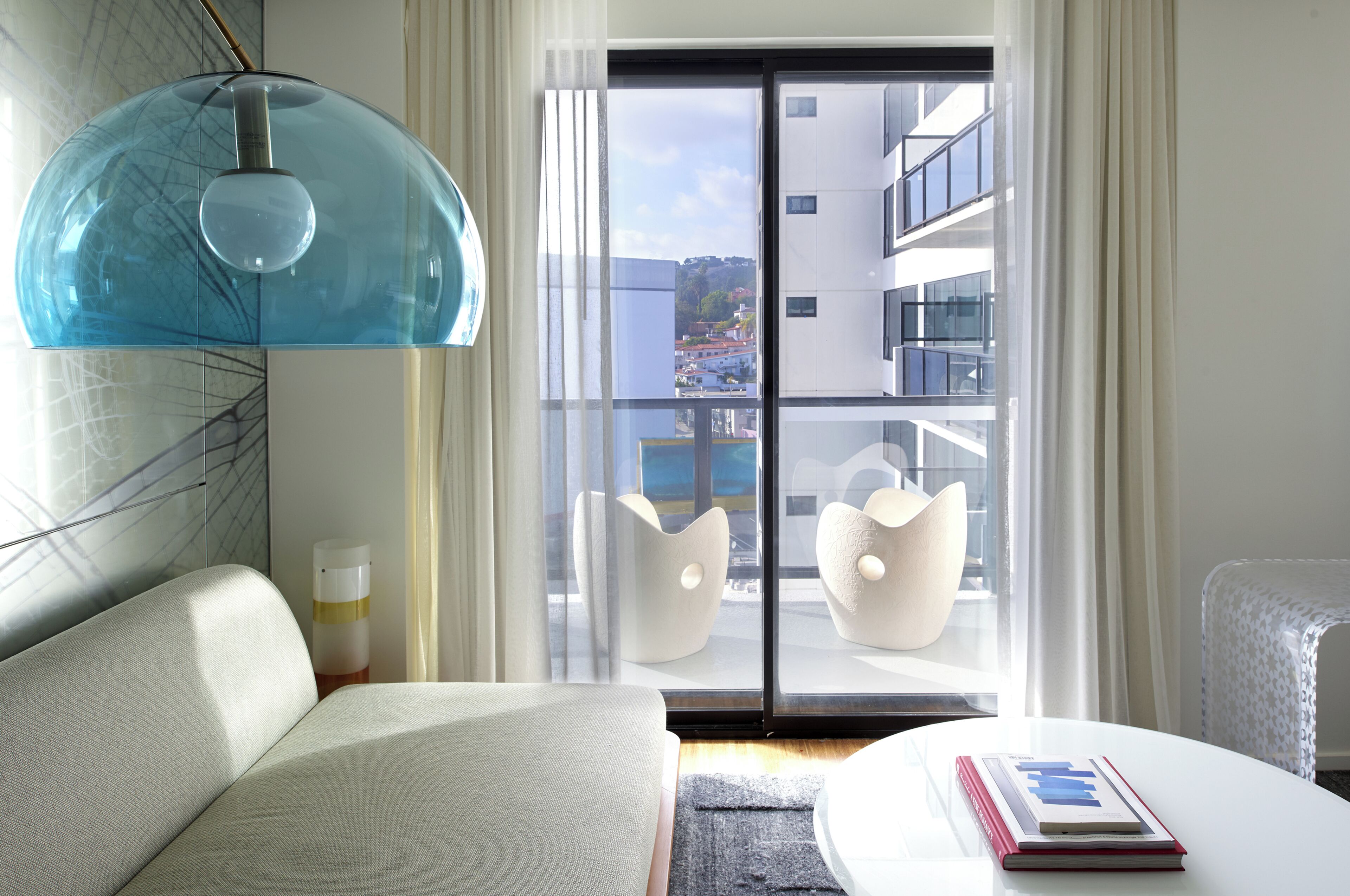 Een moderne hotelkamer met een bank, een tafel met een boek, een opvallende blauwe hanglamp en een uitzicht op een stadsgezicht door een groot raam.