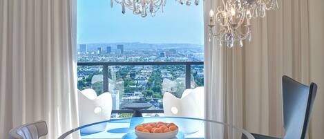 A dining area with a glass table, white chairs, a bowl of fruit, and a city view.