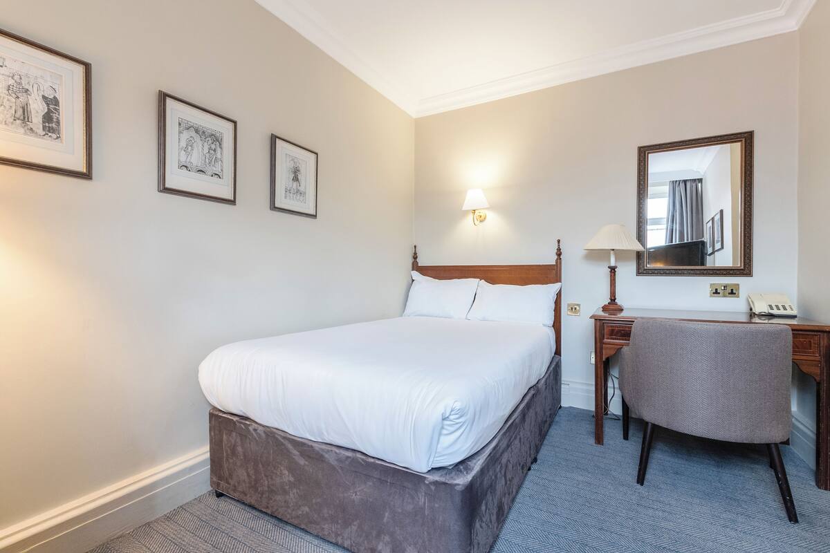 A hotel room with a bed, a desk, a chair, a lamp, and framed pictures on the wall.