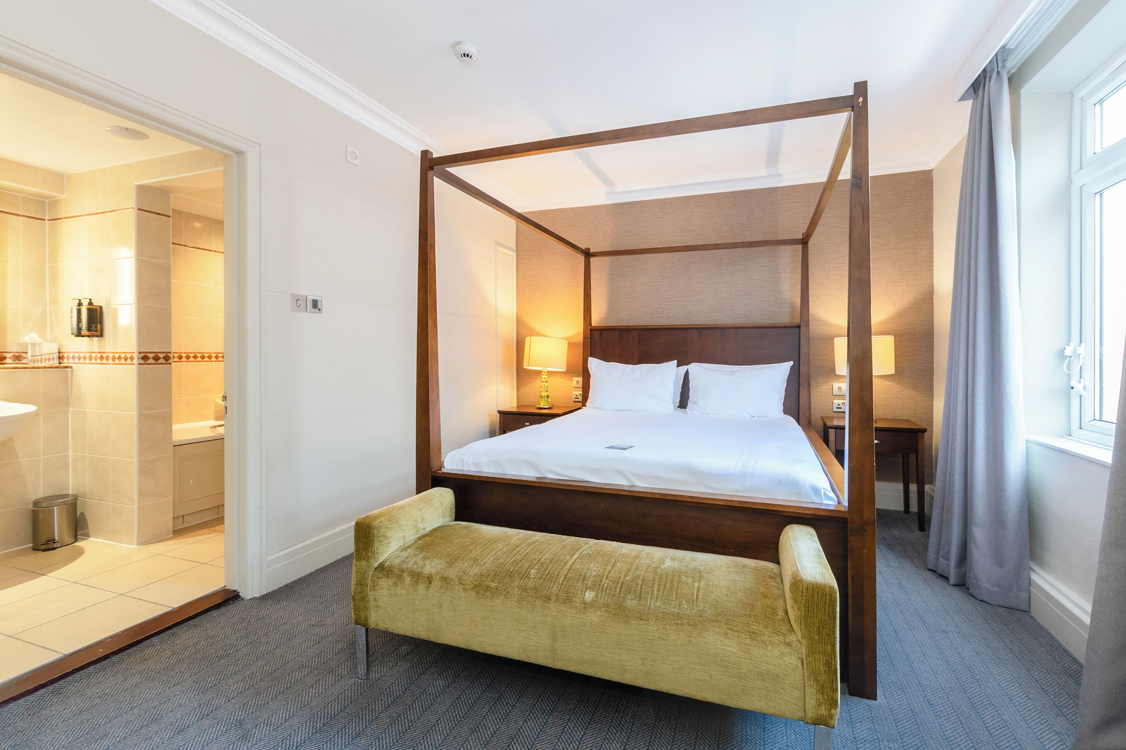 A four-poster bed with a wooden frame, a bench at the foot of the bed, and a bathroom visible through an open door.