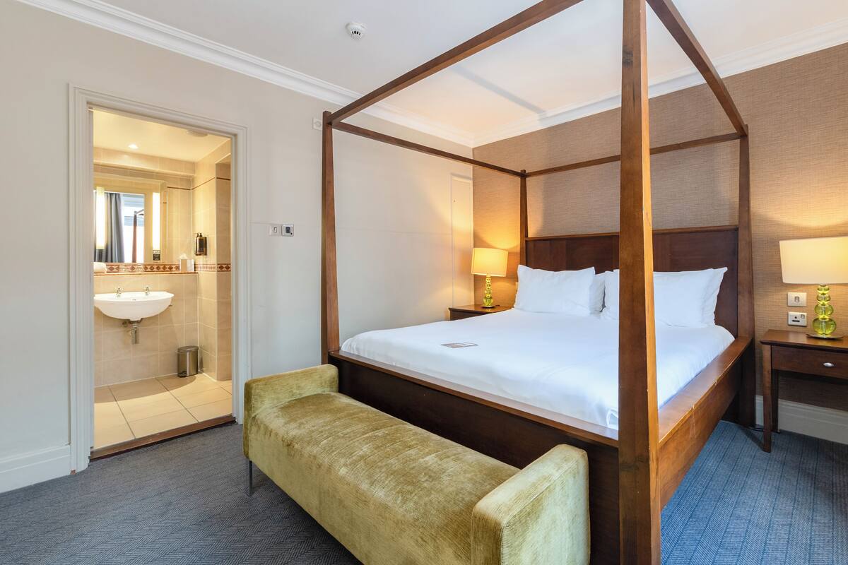 A four-poster bed with a wooden frame, a bench at the foot of the bed, a bedside table with a lamp, and a bathroom with a sink and mirror.