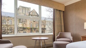 Ein Hotelzimmer mit großem Fenster und Blick auf historische Gebäude, einem runden Tisch, zwei Sesseln und einem Bett.
