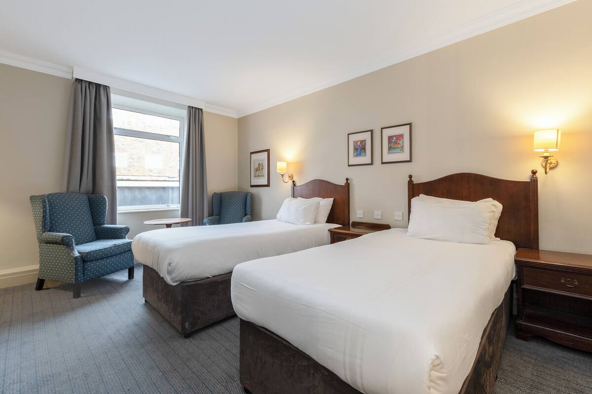 A hotel room with two beds, a chair, a nightstand, and a window with curtains.