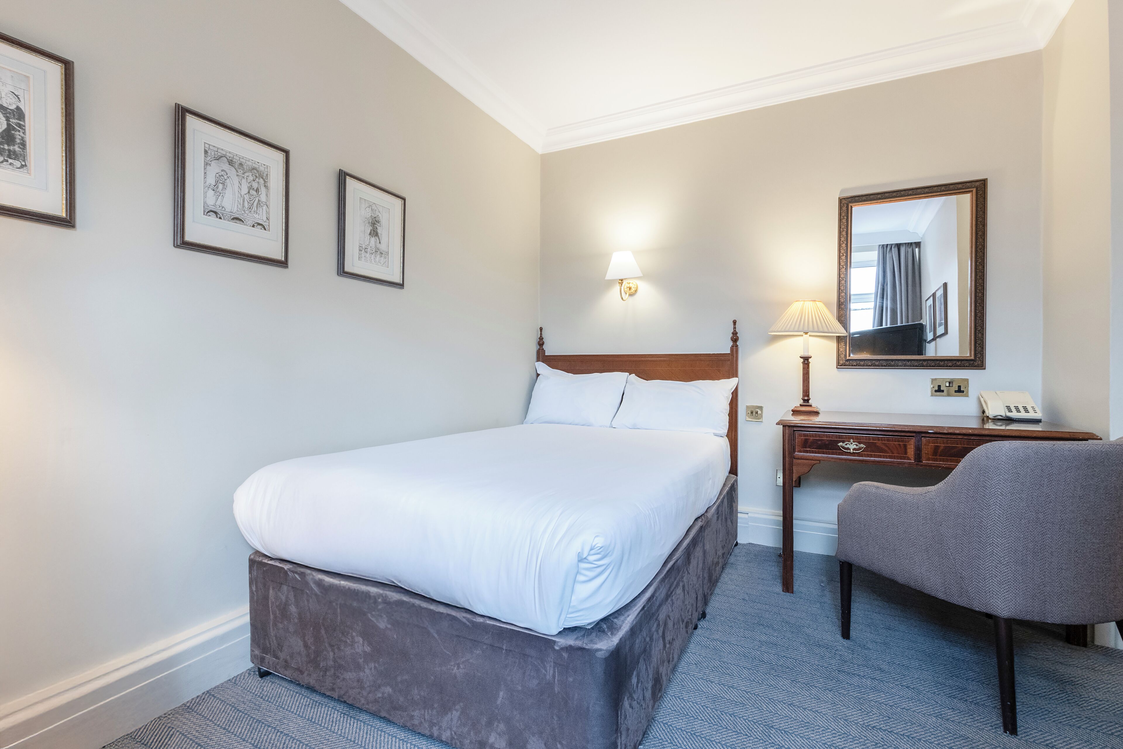 A hotel room with a large bed, a desk, a chair, and framed artwork on the wall.