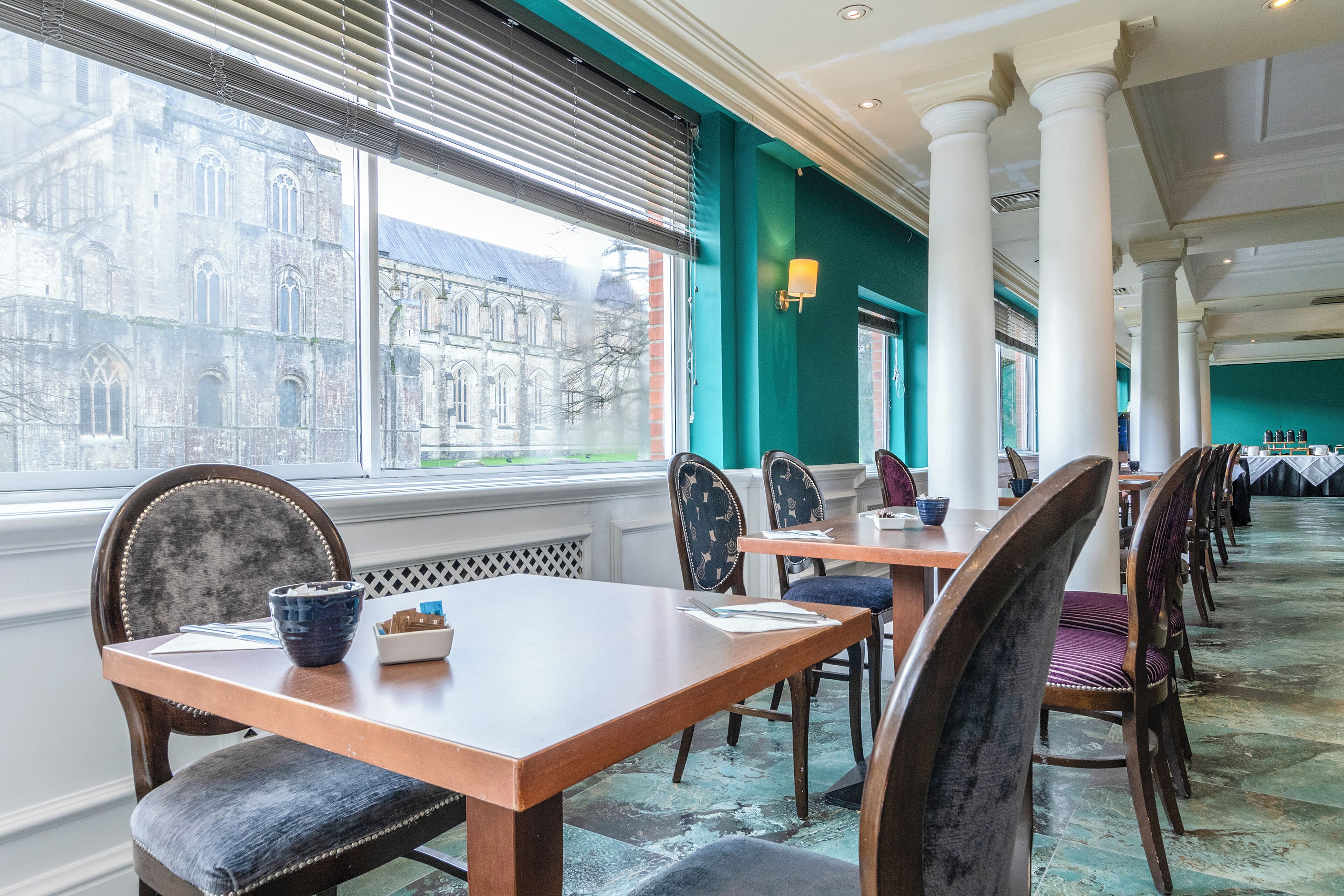 Un comedor con mesas y sillas, ventanas amplias con persianas y vista a un edificio histórico.