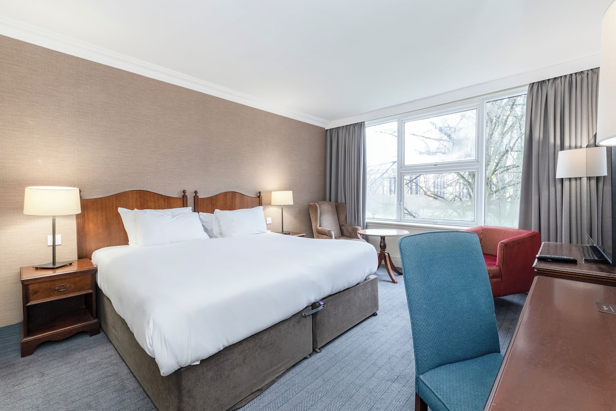 A hotel room with a large bed, two bedside tables with lamps, a desk with a chair, and a window with curtains.
