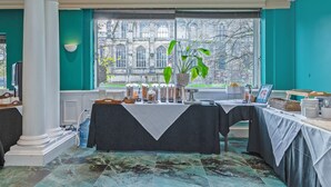 Ein Zimmer mit türkisfarbenen Wänden, ein großes Fenster mit Blick auf ein historisches Gebäude, ein Tisch mit weißer Tischdecke und eine Pflanzenarragement.