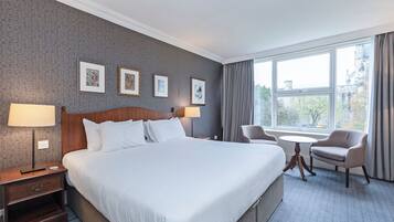 Habitación de hotel con una cama grande, dos lámparas de noche, una mesita redonda, una silla y una ventana con vista a edificios y árboles.