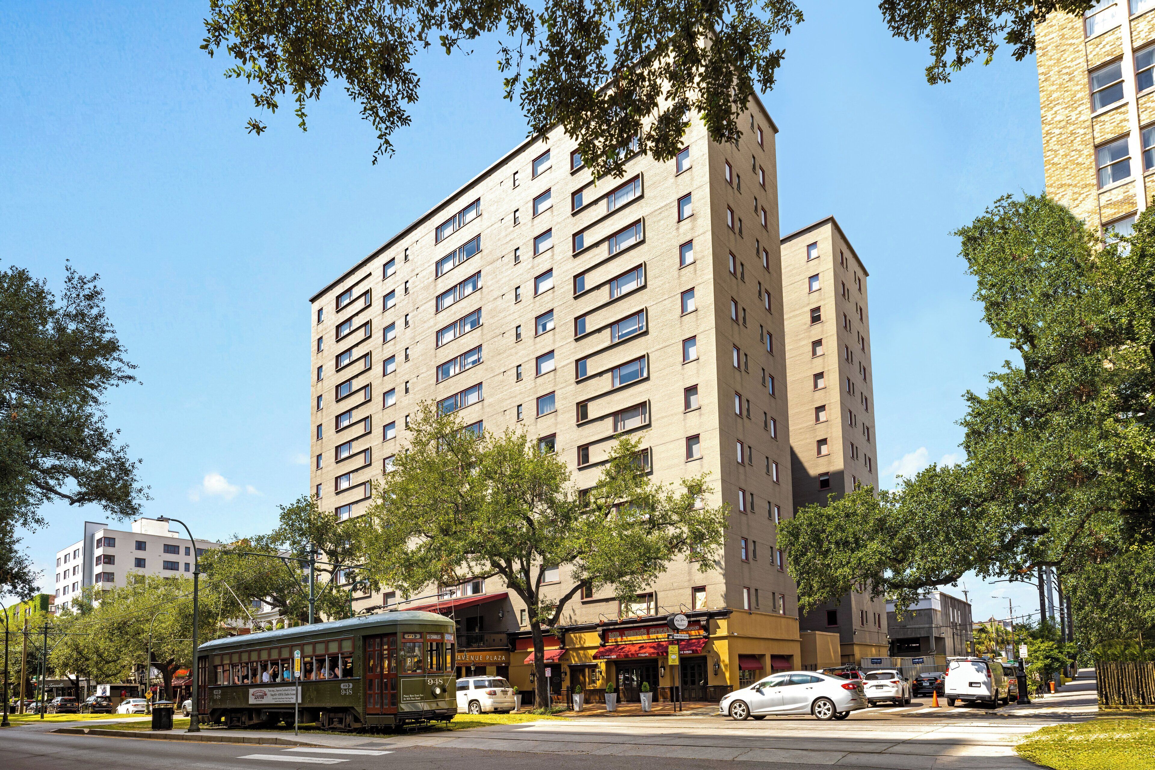 Un edificio de varios pisos con fachada plana, varias ventanas y una calle frente a él con un tranvía y autos estacionados.