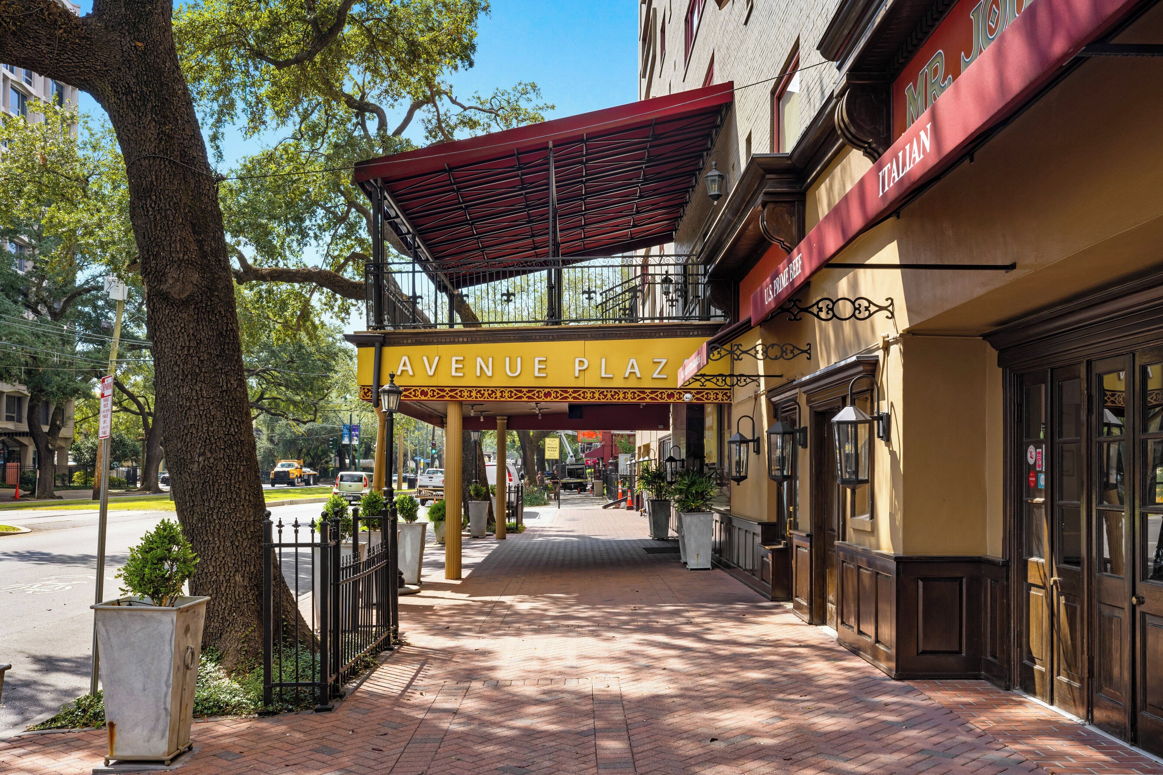 Eine überdachte Fußgängerbrücke führt zu einem Gebäude mit der Aufschrift 'AVENUE PLAZA', mit einem roten Markisenvorzelt und einem Schild für 'U.S. PRIME BEEF'.
