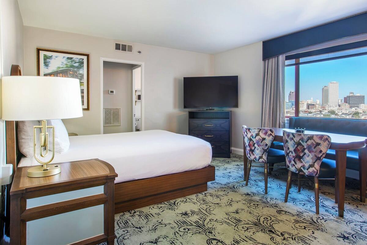 Een hotelkamer met een bed, een bureau met lamp, een televisie, een stoel, een bank en een groot raam met uitzicht op de stad.