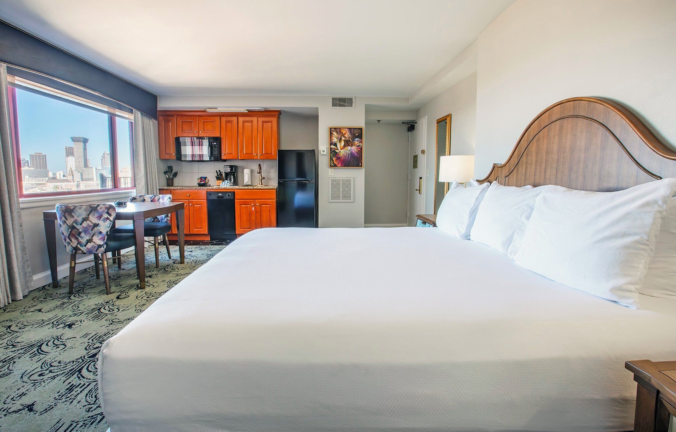 A hotel room with a large bed, a dining table with chairs, a kitchen area, and a view of the city skyline.