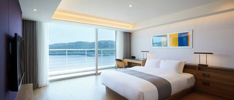 A modern hotel room with a large bed, a desk, a television, and a view of the sea.